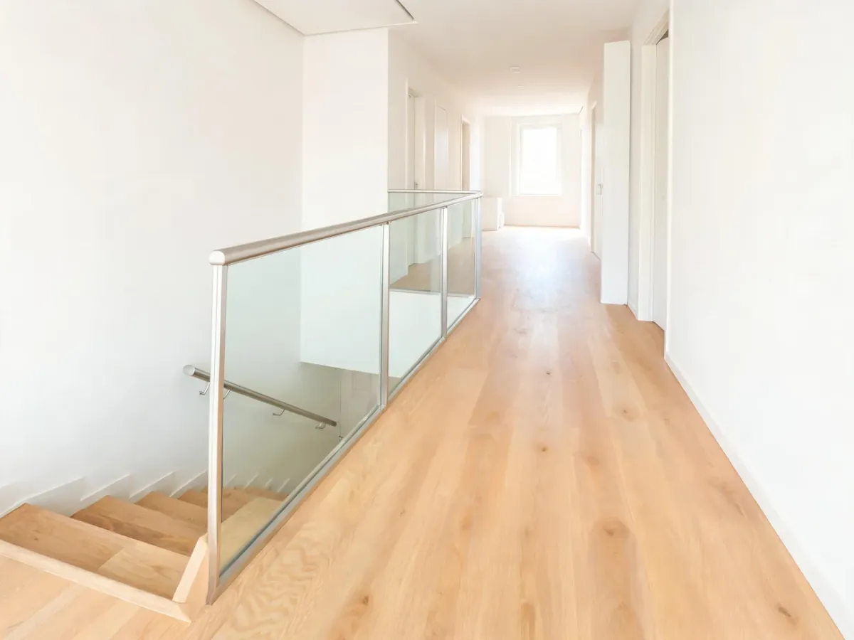 Nowoczesny korytarz z drewnianymi schodami zakończonymi bezpiecznym spocznikiem, metalowo‑szklaną balustradą i poręczami