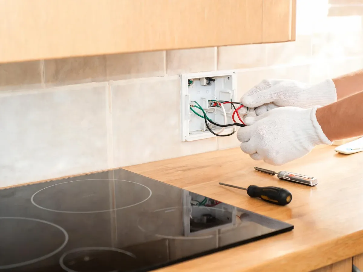 Elektryk podłącza przewody płyty indukcyjnej w puszce ściennej na tle nowoczesnej kuchni, obok leżą narzędzia i próbnik napięcia.